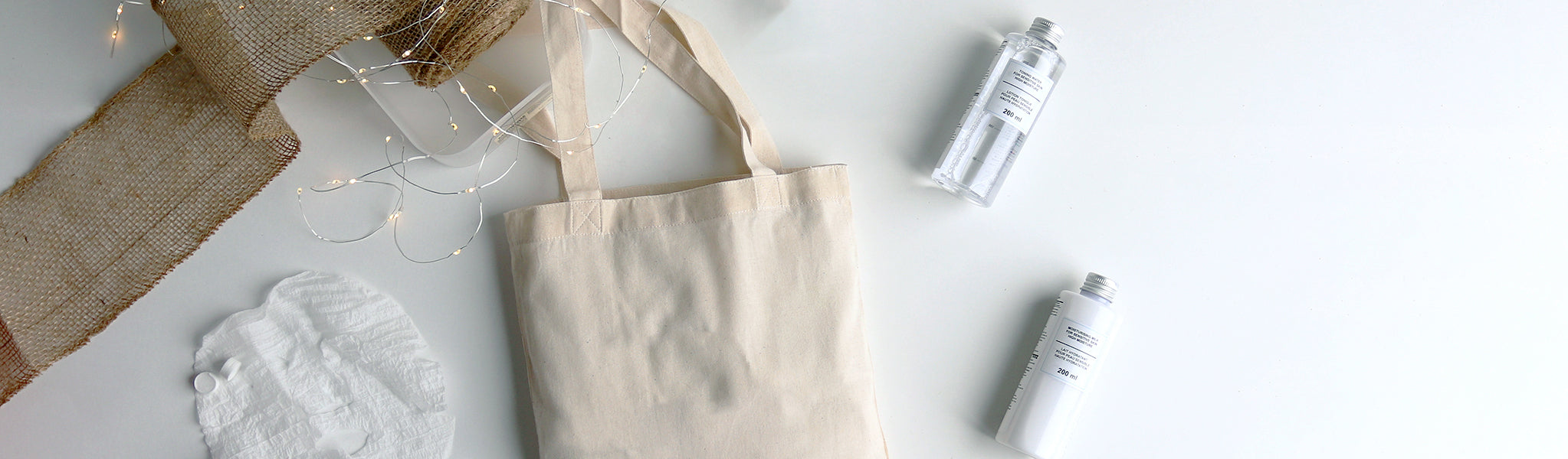 Tote bag lying flat on a white table, with two bottles of MUJI skincare products, with twinkle lights, and ribbon. 