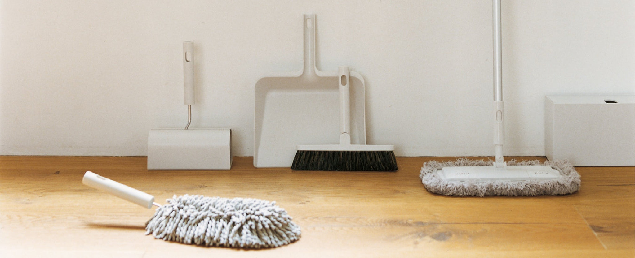 Dustpan, Broom, Mop against a white wall, handheld mop place in front on a wood floor. 
