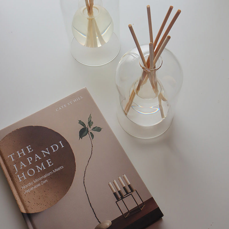 Two diffusers with wooden sticks and a book titled 'The Japandi Home' on a light surface.