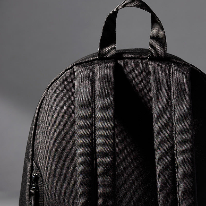 Close-up of the back of a black backpack showing padded shoulder straps and a top carry handle against a dark gray studio background.