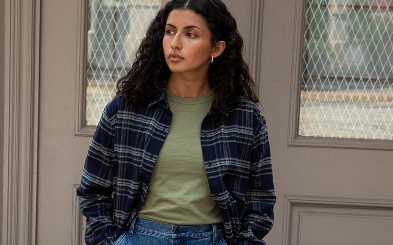 Woman wearing a blue plaid shirt over a green top and jeans, standing in front of a door.
