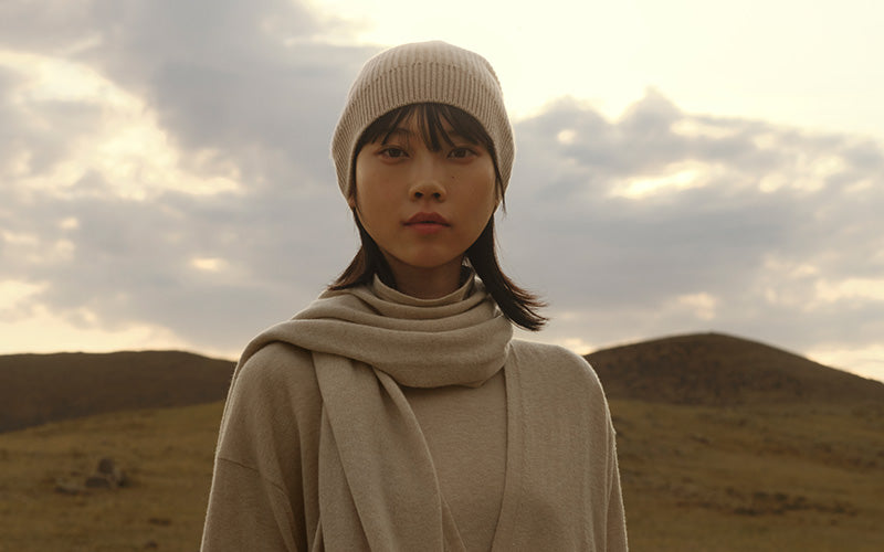 Person wearing a beige knit hat and scarf in a desert landscape with cloudy sky