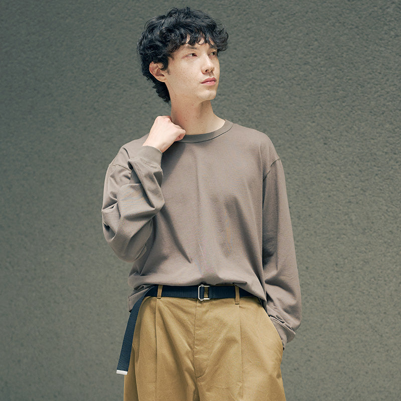 A man wearing a taupe-brown long-sleeve crewneck sweatshirt and tan trousers, standing against a textured gray wall.