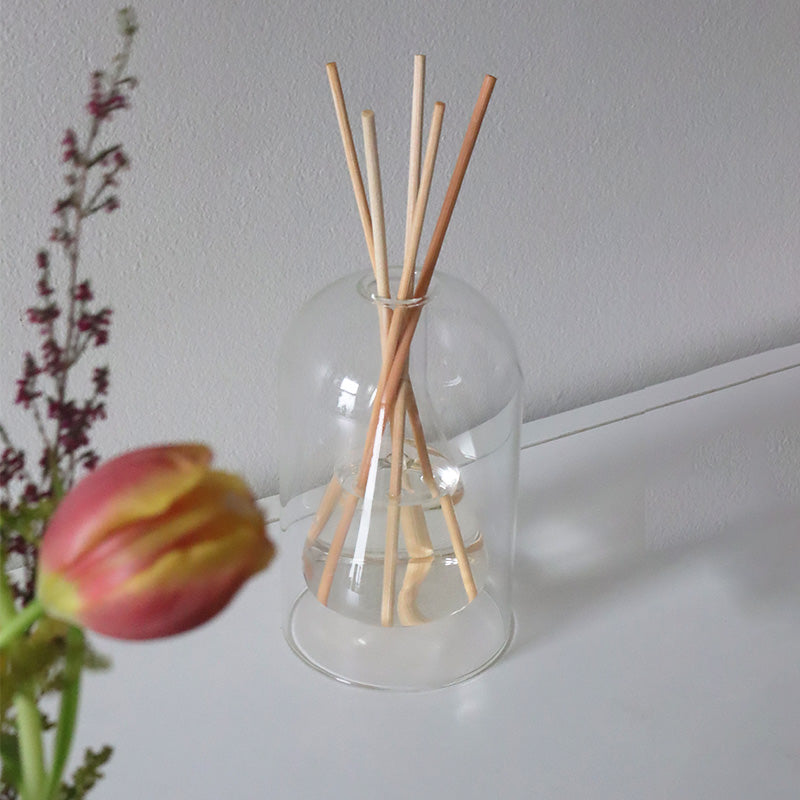 Glass Reed diffuser with wooden sticks inside on a white surface with a tulip in the foreground.