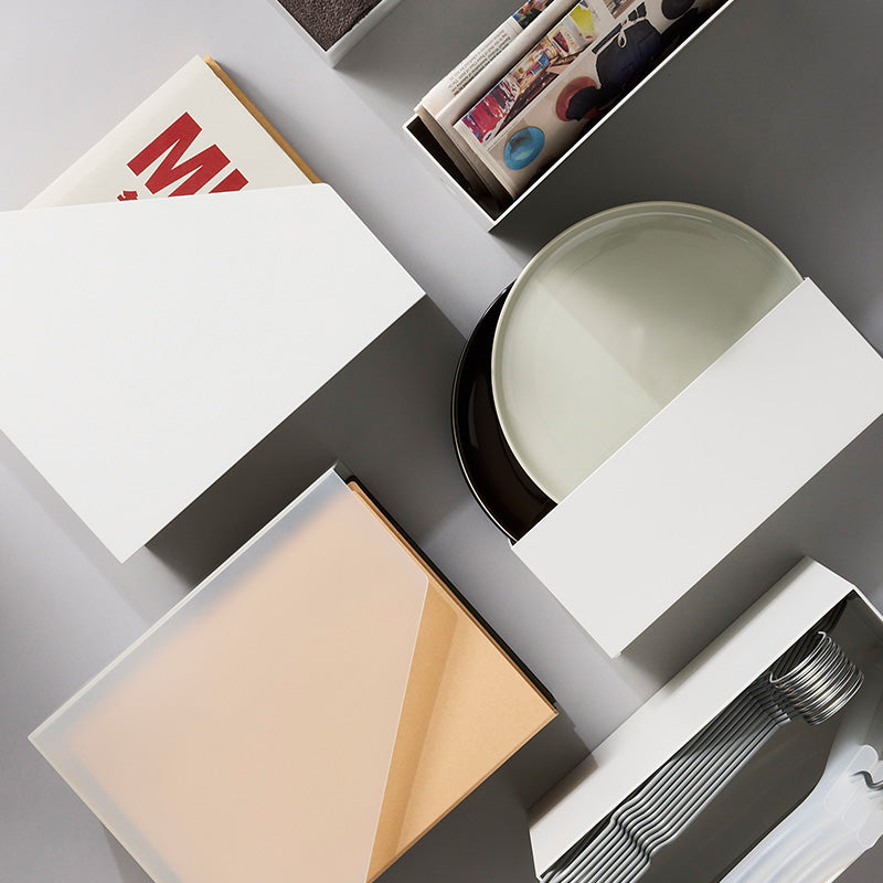 A styled overhead flat-lay of minimalist white storage boxes and containers arranged on a light gray surface, with a pale green ceramic dish and a few books or magazines partially visible.