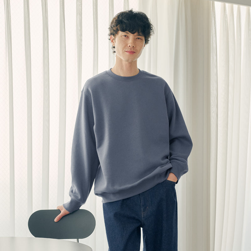 A man wearing a slate blue crewneck sweatshirt and dark blue jeans, standing in a bright room with white curtains and holding a dark chair.