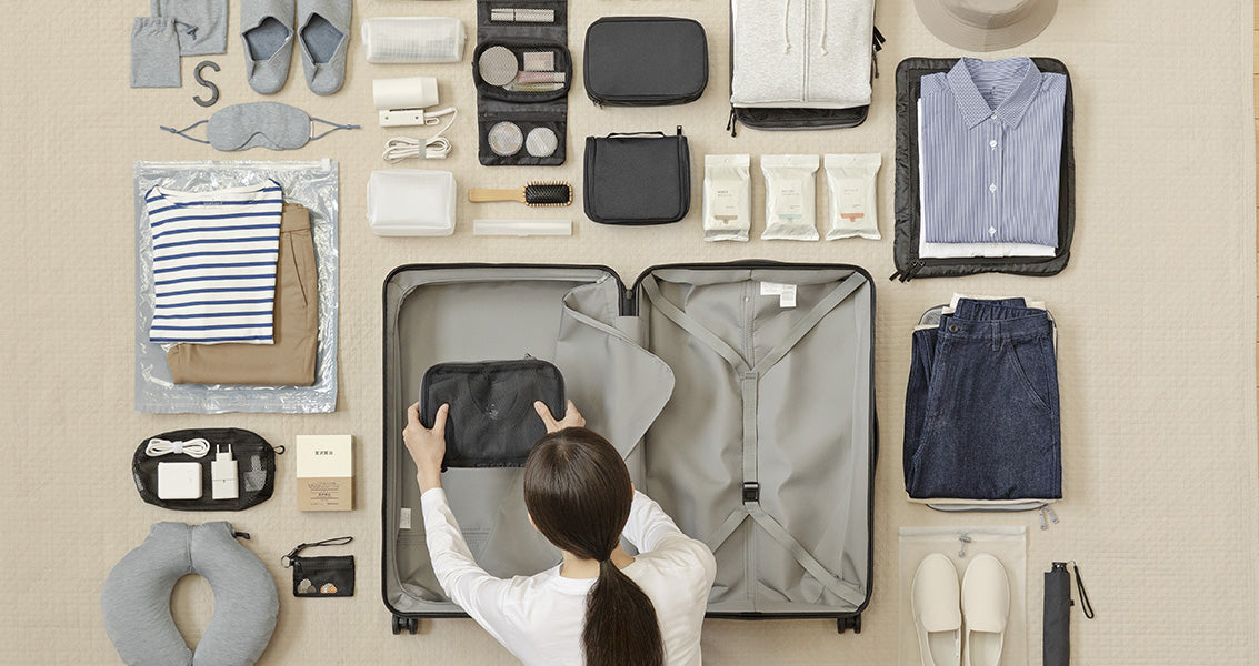 Person packing a suitcase with various travel items on a neutral background