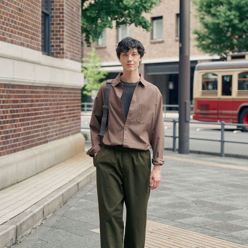 A man stands on a city sidewalk wearing a brown button-up shirt,